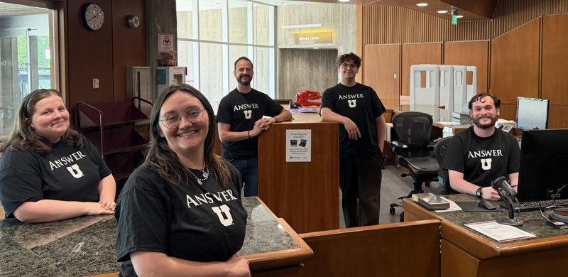 Five staff members wearing matching black “Answer U” T-shirts pose behind a service desk in a library or campus help center. The space features wood-paneled walls, service counters, office chairs, computers, and large windows that bring in natural light. One staff member stands behind a small podium, while others are positioned around the desk area. A sign on the podium references checking out equipment, and a wall sign reading “Loan Office” is visible in the background. The service area appears organized and welcoming, with workstations and customer service equipment throughout the space.