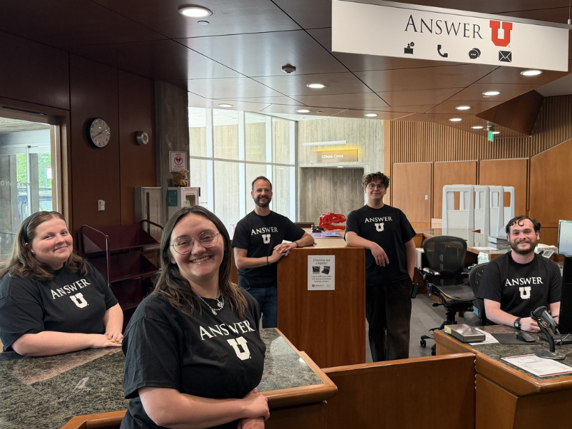Five smiling staff members in ANSWER U shirts pose at a university reception desk beneath an ANSWER U sign, creating a welcoming scene.