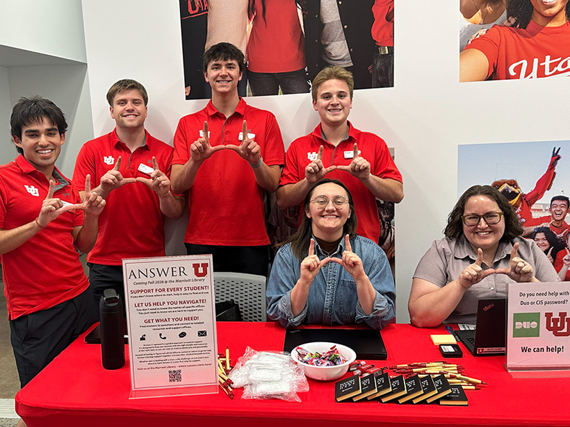 Five people sit behind a table covered with a red cloth at an informational booth. They are smiling, and three of them hold up a "U" hand sign. The table has informational materials, a bowl of candy, and two laptops. The backdrop features posters with university branding and students. The mood is welcoming and helpful.