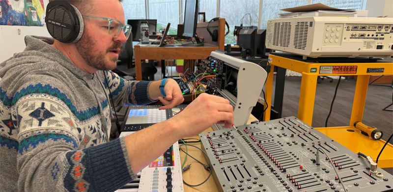 A person wearing over-ear headphones and glasses adjusts knobs and cables on a large analog synthesizer or sound-mixing console at a workbench, with a keyboard controller and electronic equipment arranged around them in a lab-like workspace.