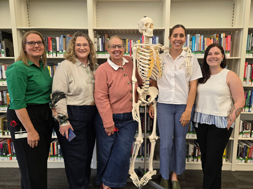 Five smiling women pose with a human skeleton model in a library, surrounded by bookshelves; a friendly, educational group photo.