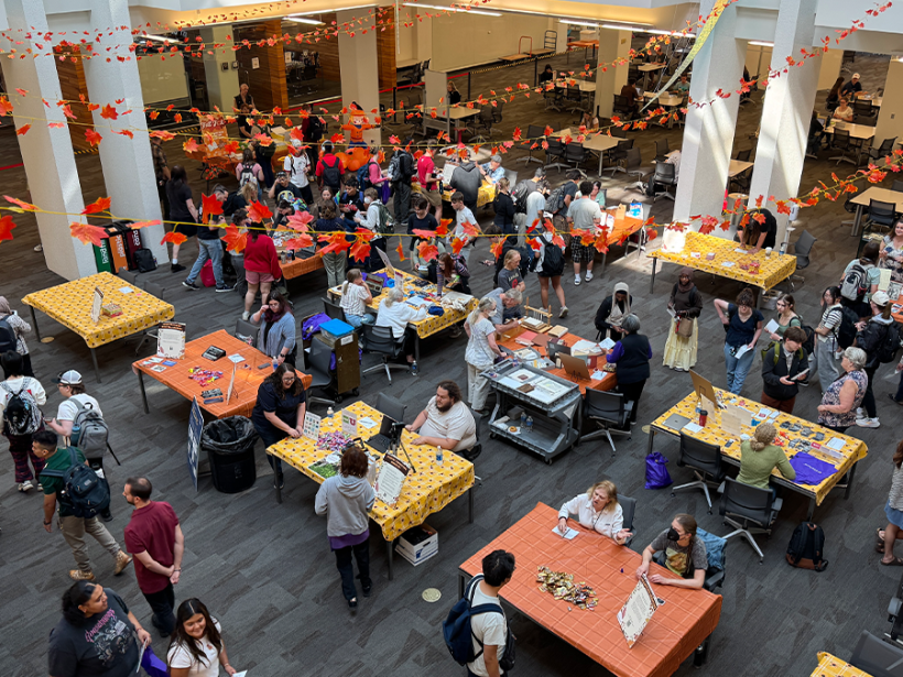 Busy indoor community fair with crowds browsing colorful vendor tables, crafts, and food; orange leaf decorations create a festive atmosphere.