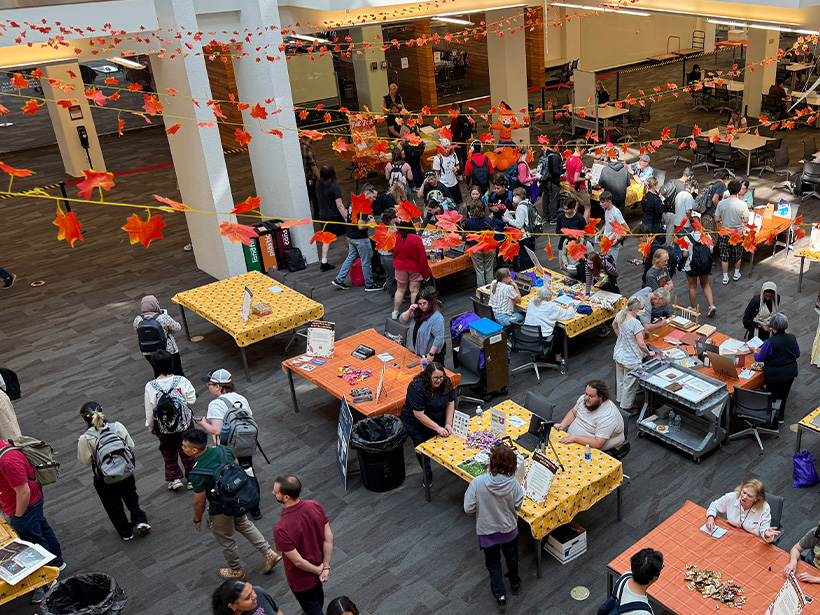 An overhead view of a busy library atrium during a large community event. Tables covered with orange and yellow tablecloths are arranged throughout the open space, displaying books, crafts, informational materials, and hands-on activities. Dozens of attendees walk between tables, browse exhibits, and speak with event organizers. Strings of red and orange paper leaves are suspended across the atrium, creating an autumn-themed decoration overhead. The space features tall white columns, open seating and study areas, and multiple service and display stations. Bright lighting and the open, multi-level layout emphasize the scale and energy of the event.