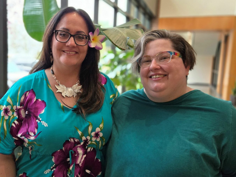 Two people smiling in a bright room. One wears glasses and a teal floral dress with a flower in her hair, the other wears a teal shirt with rainbow glasses.