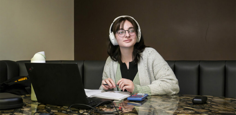 Young woman with glasses and headphones sits at a marble table with a laptop, notebook and calculator.