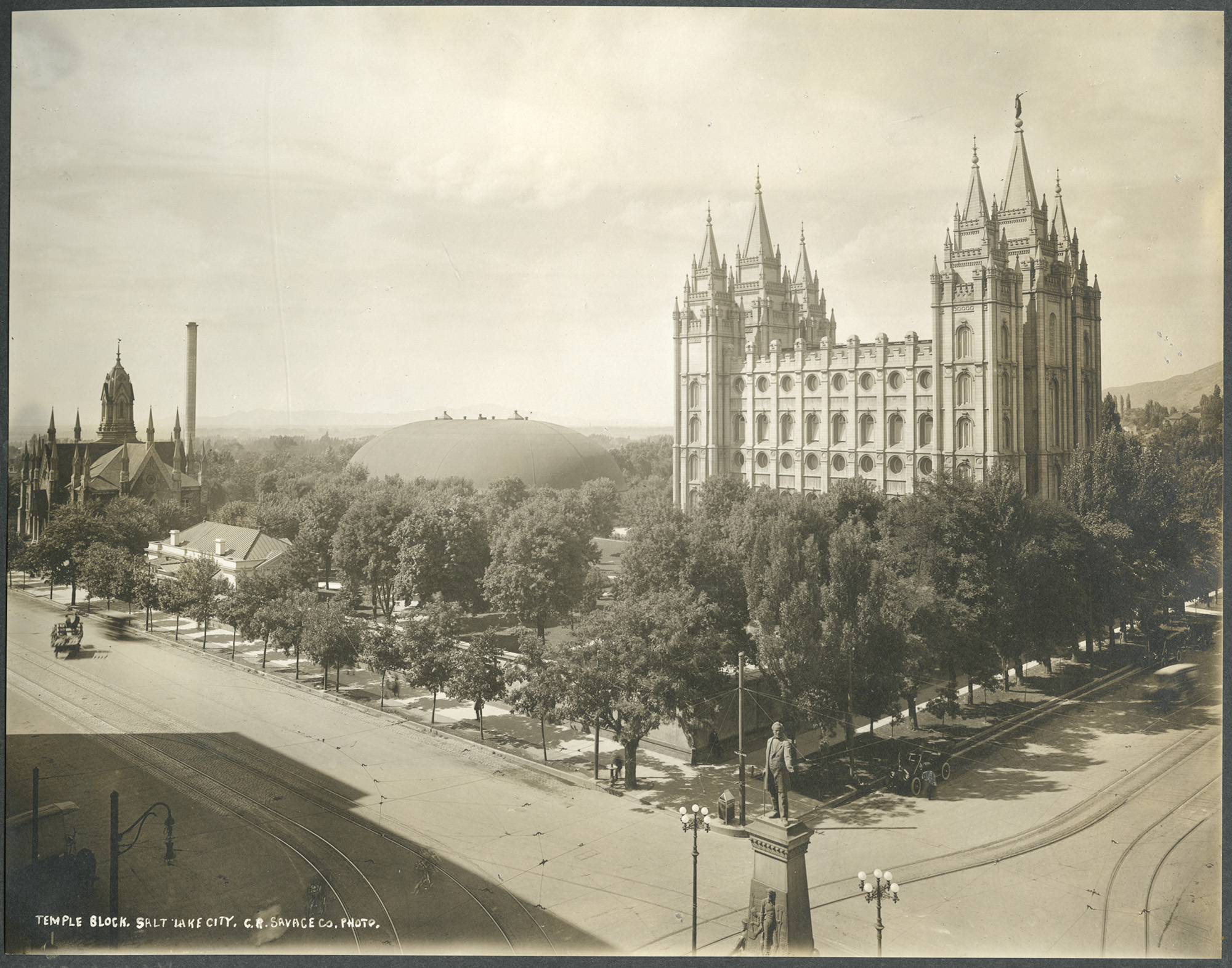 Historic black-and-white view of Salt Lake Temple with ornate spires, tree-lined streets, horse-drawn vehicles, and pedestrians.