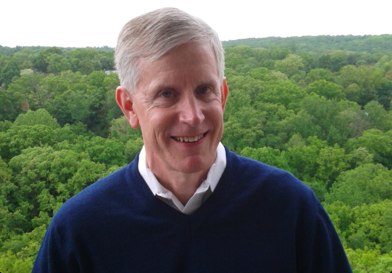 val, a white man with white hair and a blue sweater, standing in front of a canopy of trees