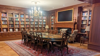 elegant room with a dining table and 10 chairs with a chandelier overhead and two armchairs surrounded by shelves of books